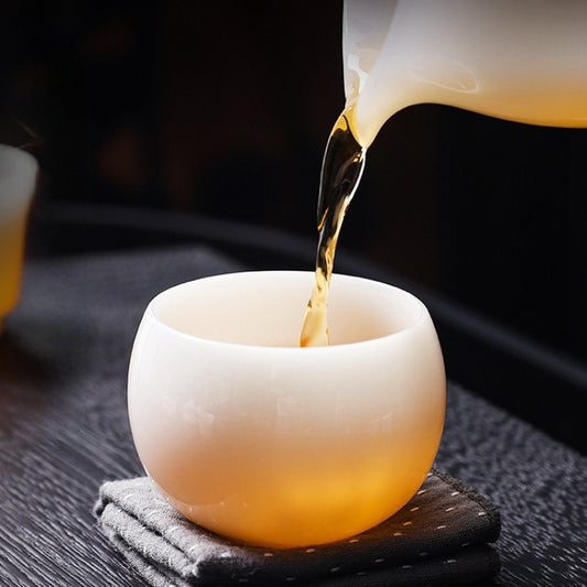 A small round white ceramic tea cup sits on a dark coaster while amber tea is being poured from a matching vessel into it.