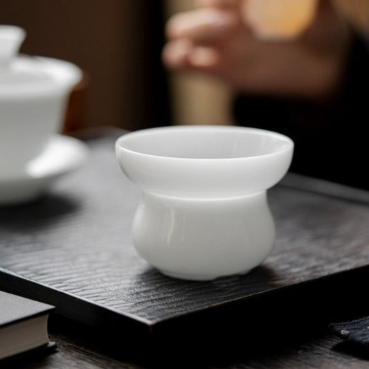 A small white ceramic cup with a wide rim and rounded base, placed on a dark wooden tray in a softly blurred tea-setting scene.