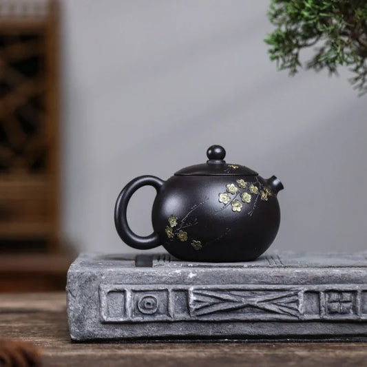 A black ceramic teapot with gold floral detailing, resting on a gray decorative stone block on a wooden table.