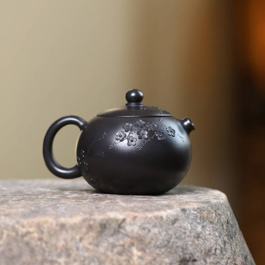 A small black ceramic teapot with floral engravings, resting on a textured stone surface against a softly blurred beige background.
