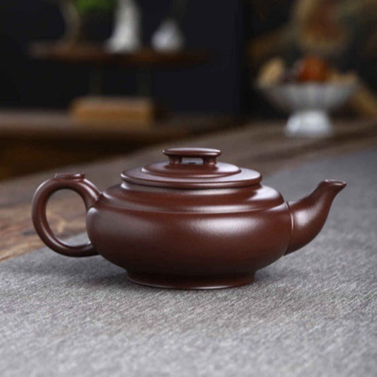 A classic Yixing clay teapot in a rich reddish-brown color, resting on a gray textured mat with a softly blurred tea house background.