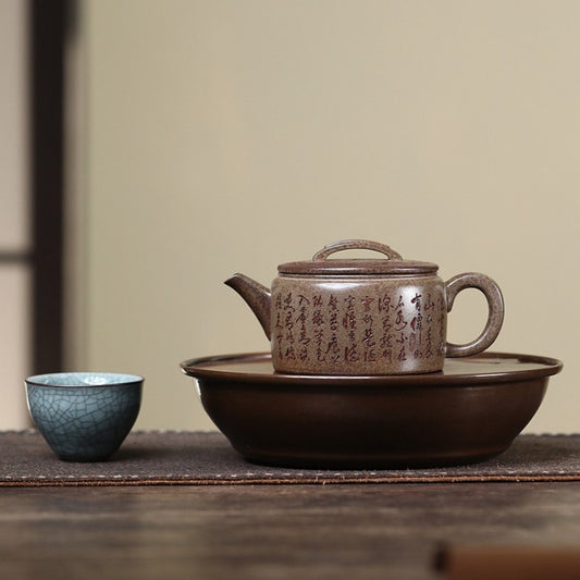 A brown ceramic teapot with Chinese calligraphy engraved on its side, resting on a matching brown teapot tray, with a small blue-green crackled celadon cup beside it on a wooden table.