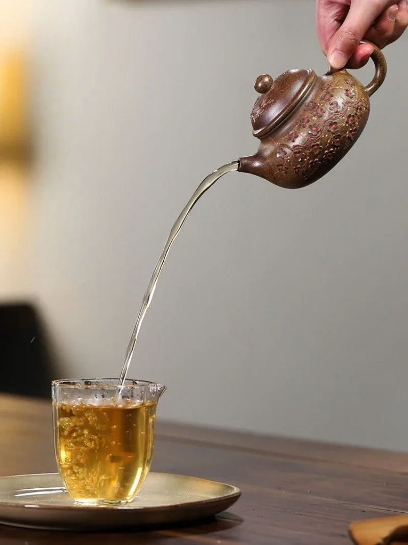 A hand holds a small brown ceramic teapot pouring amber tea into a clear glass cup on a saucer, set on a wooden table against a softly blurred neutral background.
