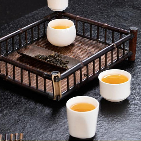 A bamboo tea tray with a woven mat and metal corner brackets, holding a small bowl of loose tea leaves and three white ceramic cups filled with amber tea, set on a dark textured surface.

