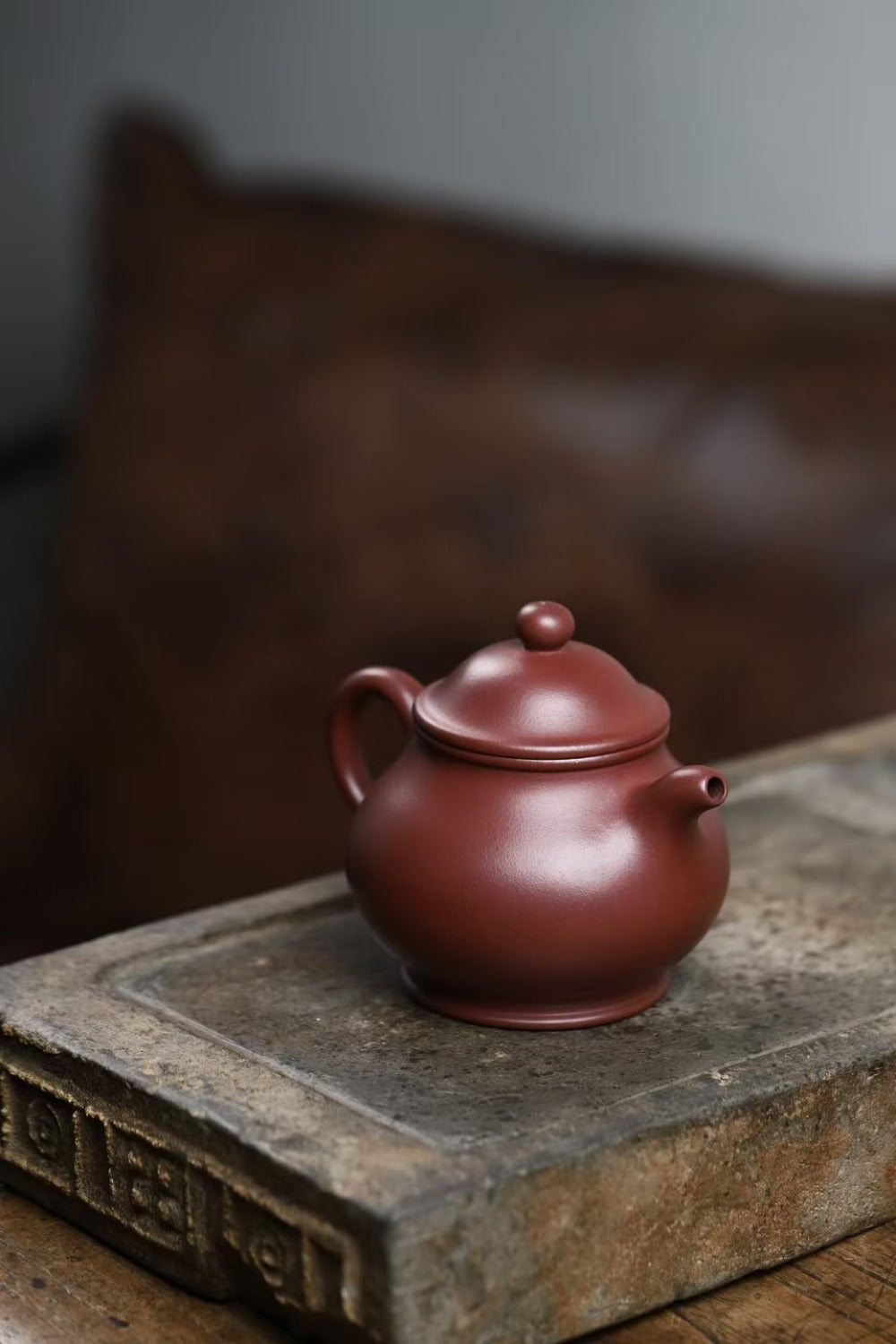 A small round reddish-brown ceramic teapot with a lid, placed on a textured stone slab with a blurred dark brown background.

