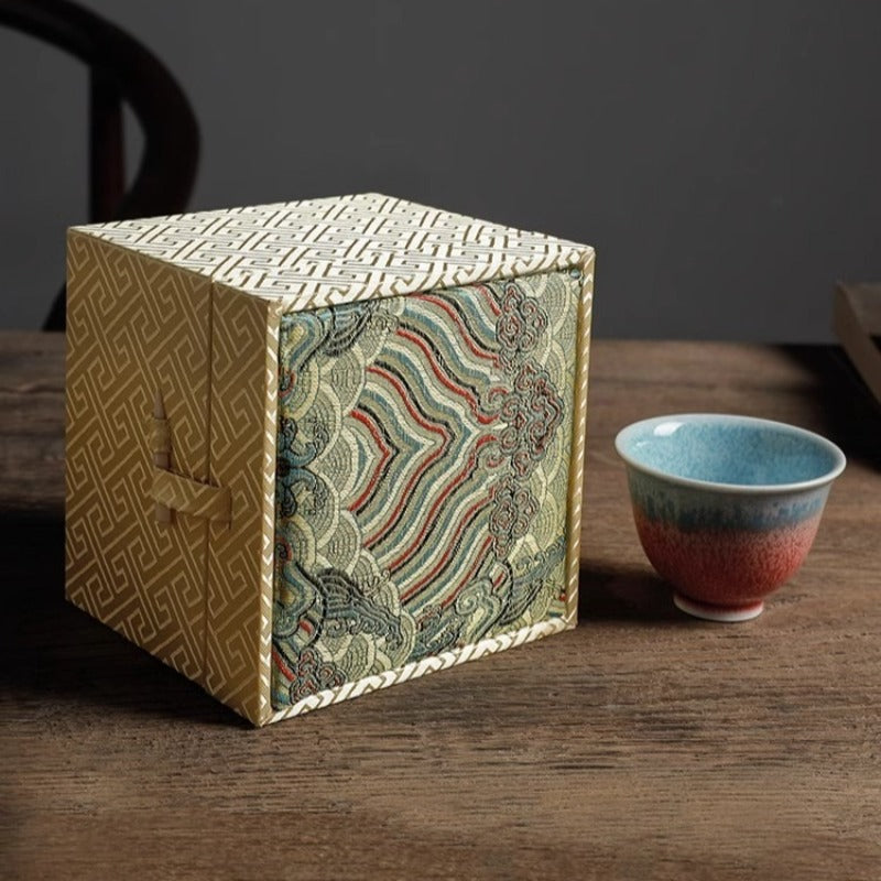 ornate cubic gift boxes with traditional East Asian-inspired designs on a wooden table: a yellow box with geometric patterns and a white ribbon clasp in the front left, a red box with similar geometric patterns and ribbon clasp in the front right, and a larger blue-gray box stacked on top featuring intricate wave and cloud motifs in teal, purple, and gold tones. In the blurred background, a traditional chair and teapot are visible.