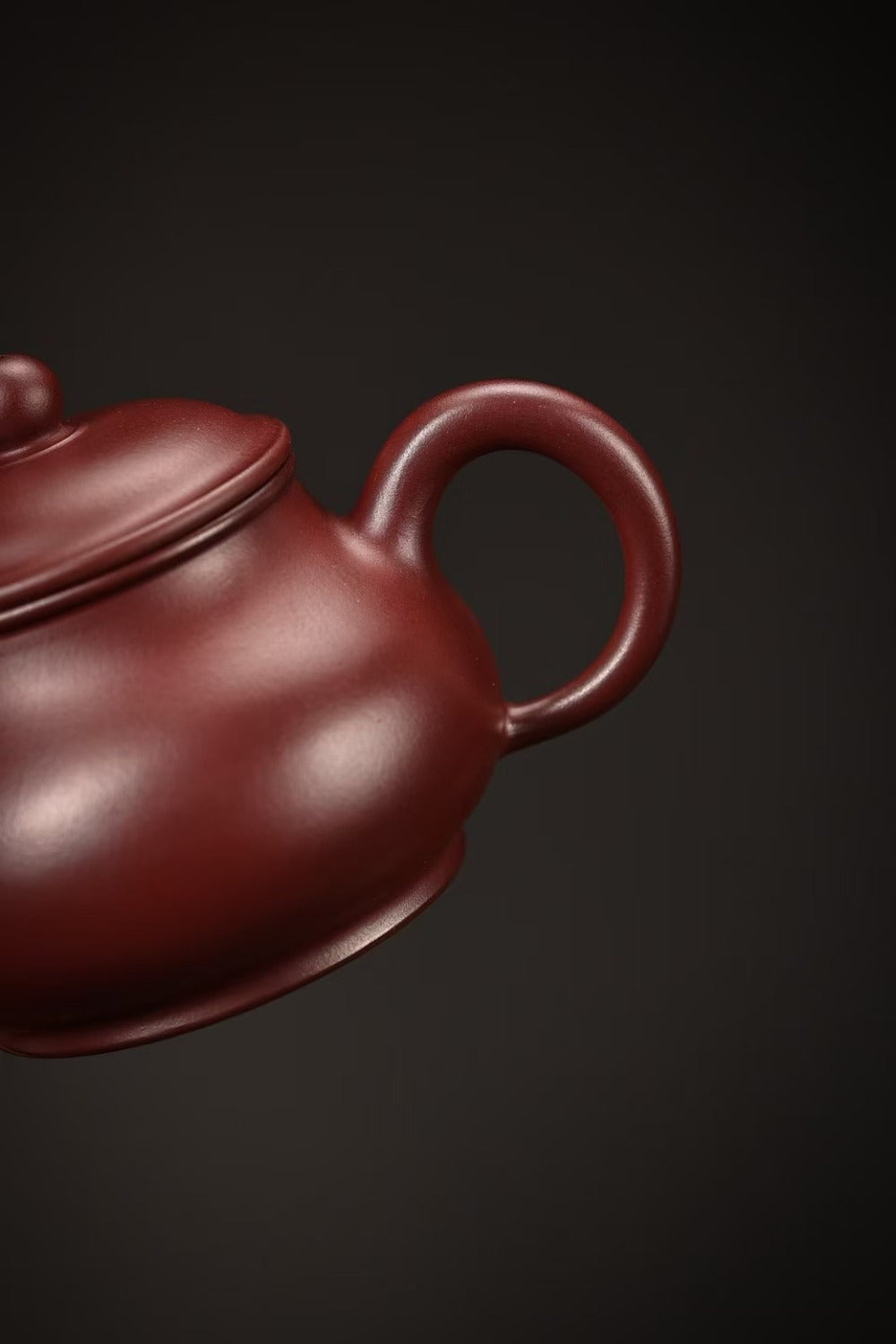 A close-up of a traditional Chinese Yixing clay teapot in a deep reddish-brown color, showing the lid, handle, and curved body against a dark background.

