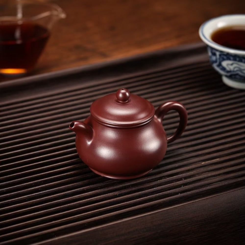 A classic Chinese Yixing clay teapot in a reddish-brown color, resting on a dark bamboo tea tray with a blurred background showing tea cups.

