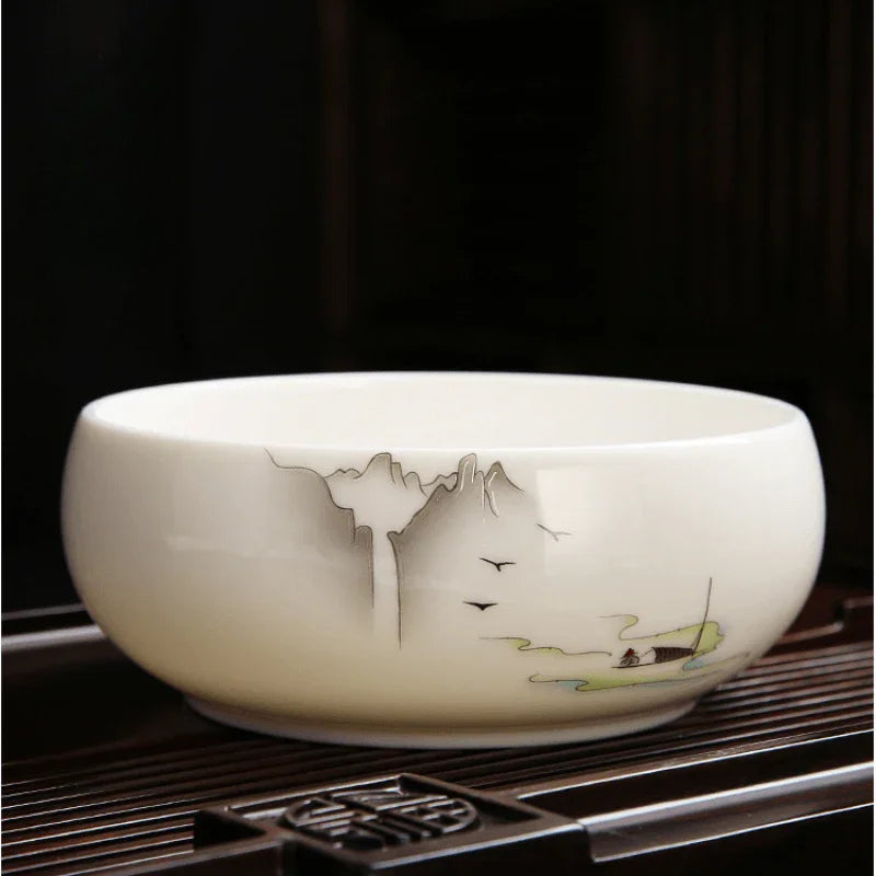 A round white ceramic bowl with hand-painted traditional Chinese landscape motifs, including mountains, birds, and a small boat, resting on a dark bamboo tea tray.

