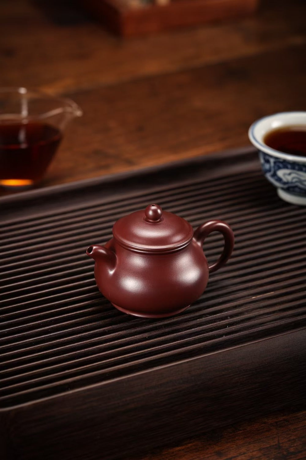 A small dark reddish-brown ceramic teapot sits on a dark wooden tea tray, with a glass cup of tea and a blue-and-white teacup blurred in the background on a wooden table.

