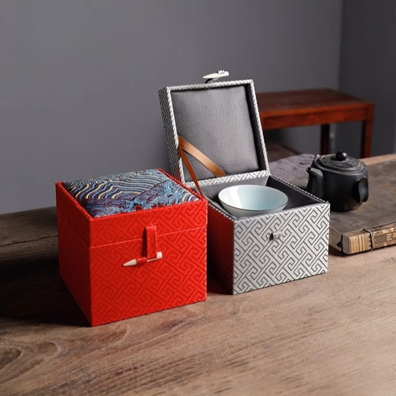 Three ornate cubic gift boxes with traditional East Asian-inspired designs on a wooden table: a yellow box with geometric patterns and a white ribbon clasp in the front left, a red box with similar geometric patterns and ribbon clasp in the front right, and a larger blue-gray box stacked on top featuring intricate wave and cloud motifs in teal, purple, and gold tones. In the blurred background, a traditional chair and teapot are visible.