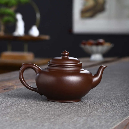 A classic dark brown clay teapot with a matching lid, resting on a gray textured tea mat in a styled tea room setting.

