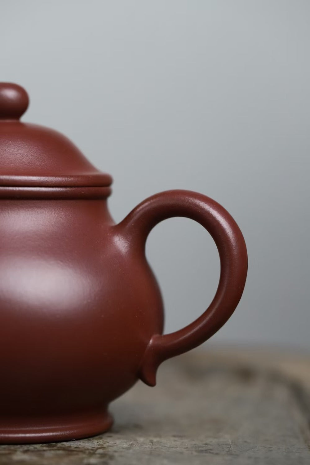 A close-up of a traditional Chinese clay teapot in a reddish-brown color, showing the side of the body, lid, and handle, resting on a rustic wooden surface against a neutral gray background.

