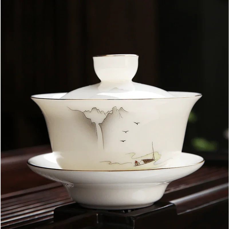 A white porcelain gaiwan set with a lid and saucer, featuring delicate gold and gray hand-painted mountain and boat designs, displayed on a dark wooden stand.

