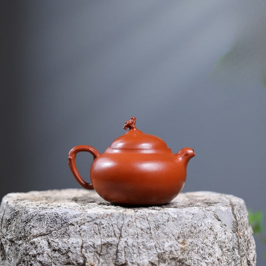 A classic Chinese clay teapot in a reddish-brown terracotta color, resting on a textured stone block against a soft, blurred gray background.

