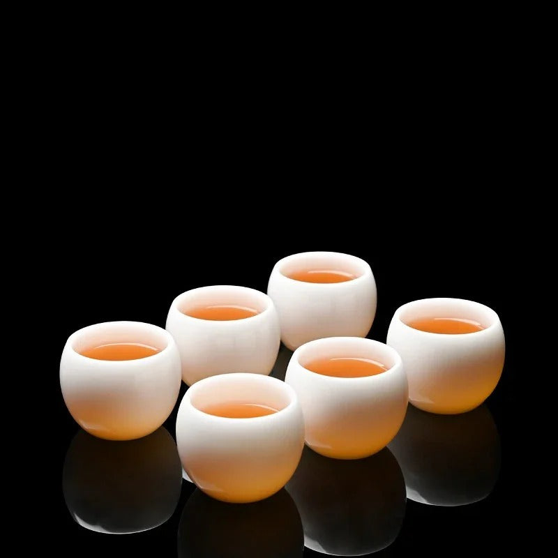 Six small white ceramic tea cups arranged in a cluster on a glossy black reflective surface, each filled with amber-colored tea against a solid black background.

