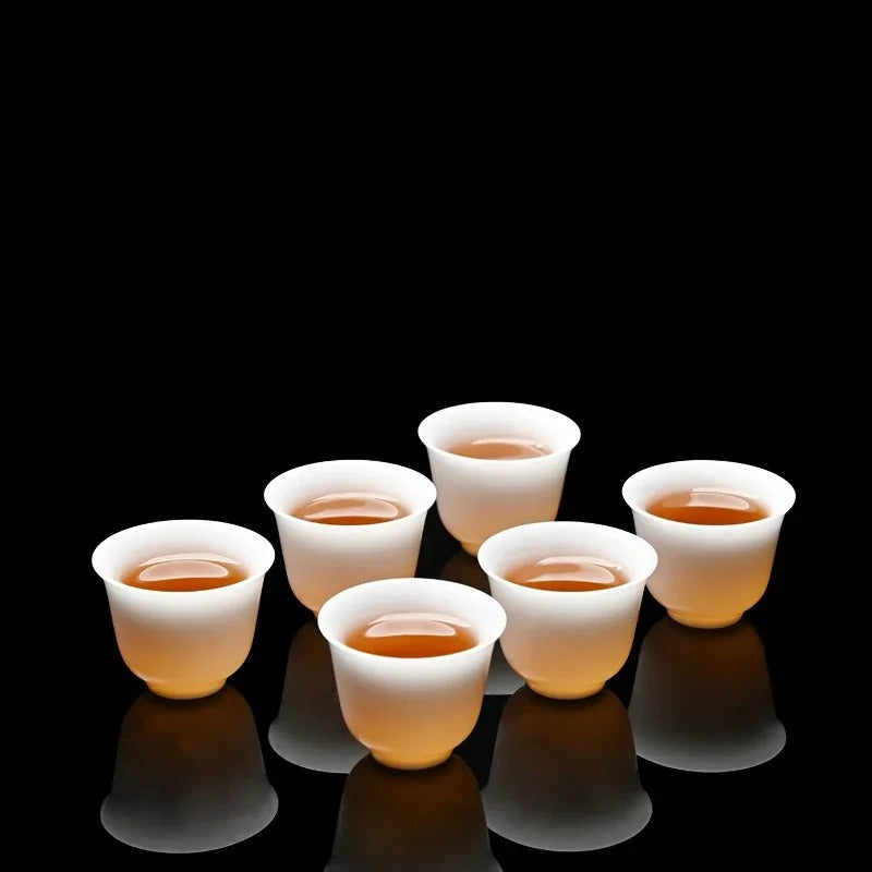 Five small translucent white cups arranged on a glossy black reflective surface, each filled with amber tea, against a solid black background.


