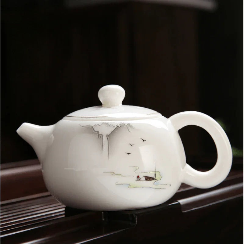 A white ceramic teapot with delicate hand-painted mountain and boat illustrations, resting on a dark wooden surface against a dark background.


