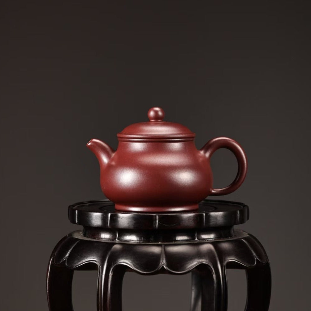 A classic Chinese Yixing Zisha teapot in a deep reddish-brown clay color, displayed on a black wooden stand against a dark gray background.

