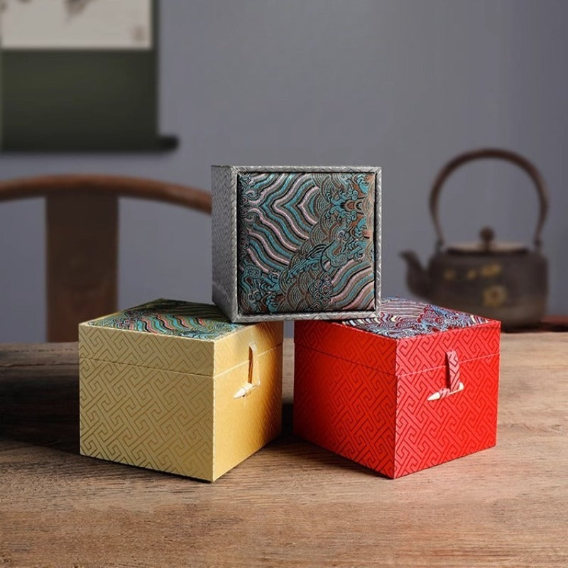 Three ornate cubic gift boxes with traditional East Asian-inspired designs on a wooden table: a yellow box with geometric patterns and a white ribbon clasp in the front left, a red box with similar geometric patterns and ribbon clasp in the front right, and a larger blue-gray box stacked on top featuring intricate wave and cloud motifs in teal, purple, and gold tones. In the blurred background, a traditional chair and teapot are visible.