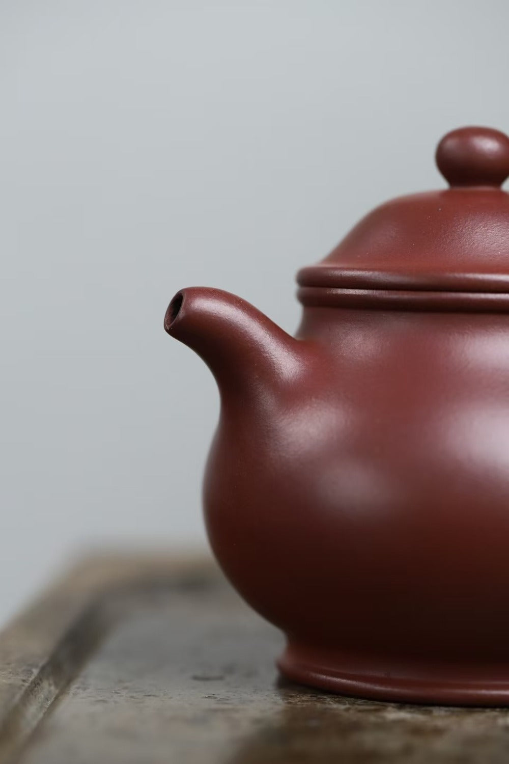 A close-up of a classic Chinese Yixing clay teapot in a reddish-brown color, resting on a rustic wooden surface against a soft gray background.

