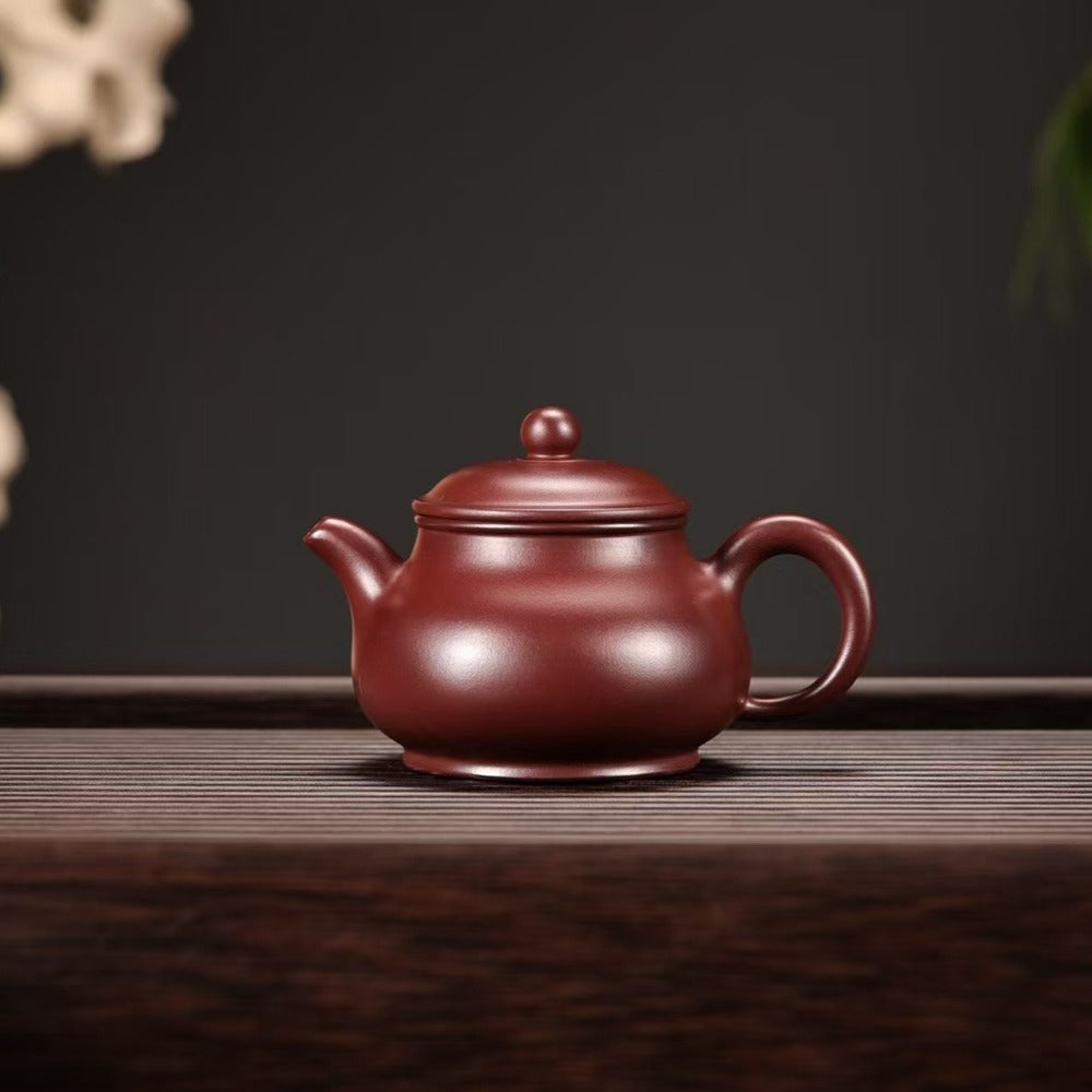 A classic Yixing clay teapot in a deep reddish-brown color, sitting on a dark wooden table against a dark gray background with subtle decorative elements in the corners.


