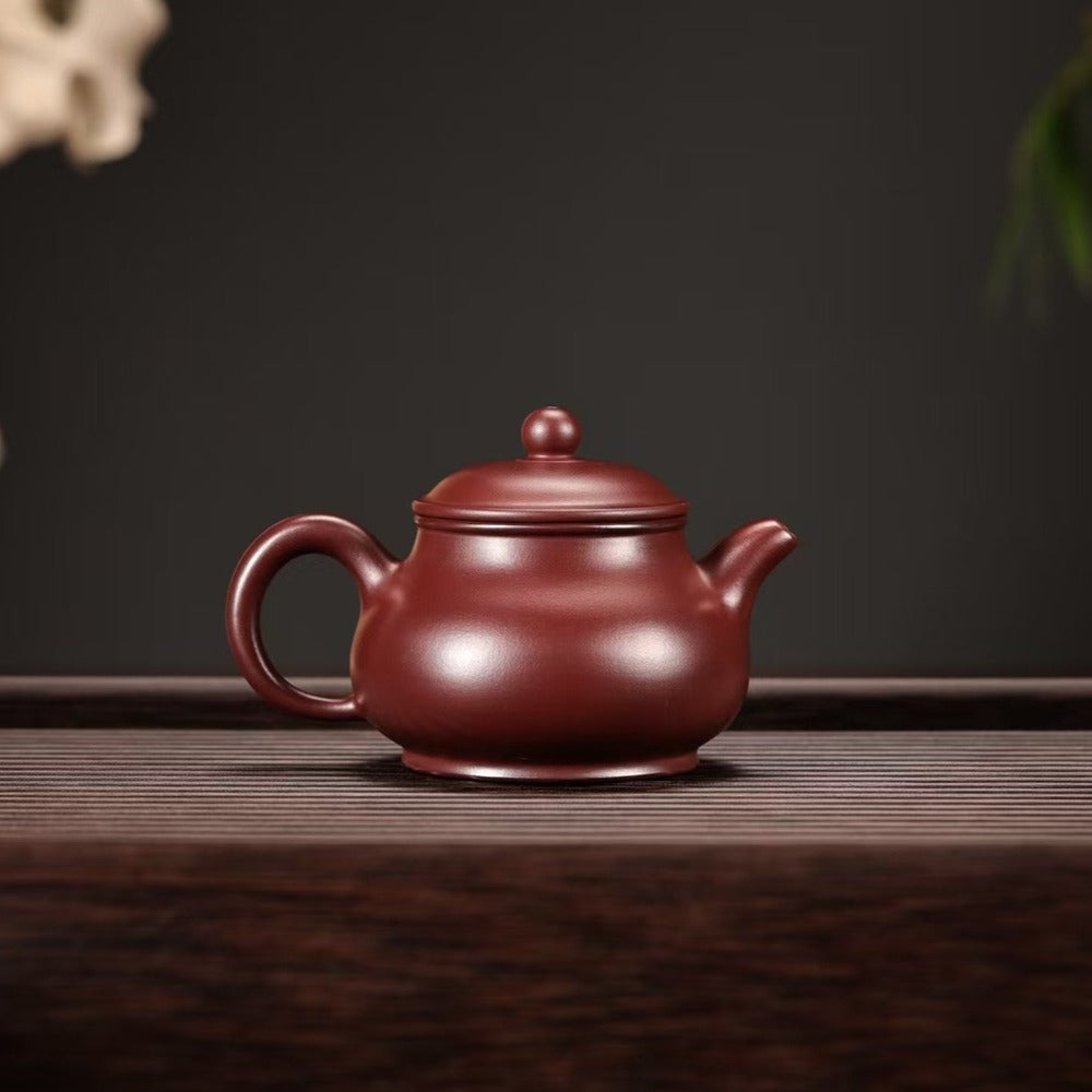 A classic Chinese Yixing Zisha teapot in a deep reddish-brown clay finish, resting on a dark wooden table against a dark gray background.

