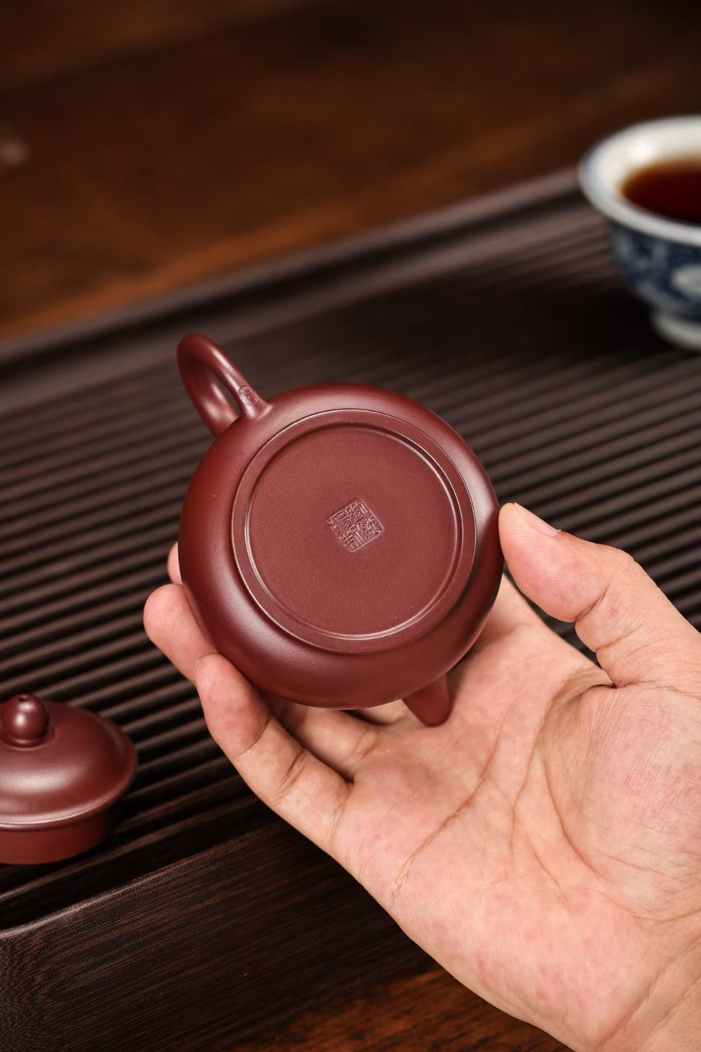 A hand holding the bottom of a small round reddish-brown ceramic teapot or vessel, with a matching lid nearby on a dark wooden tray and a cup in the blurred background.

