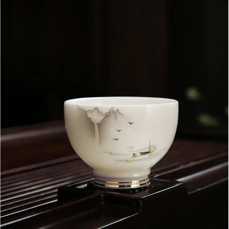 A white ceramic tea bowl with delicate hand-painted mountain and boat motifs, displayed on a dark wooden stand against a dark background.


