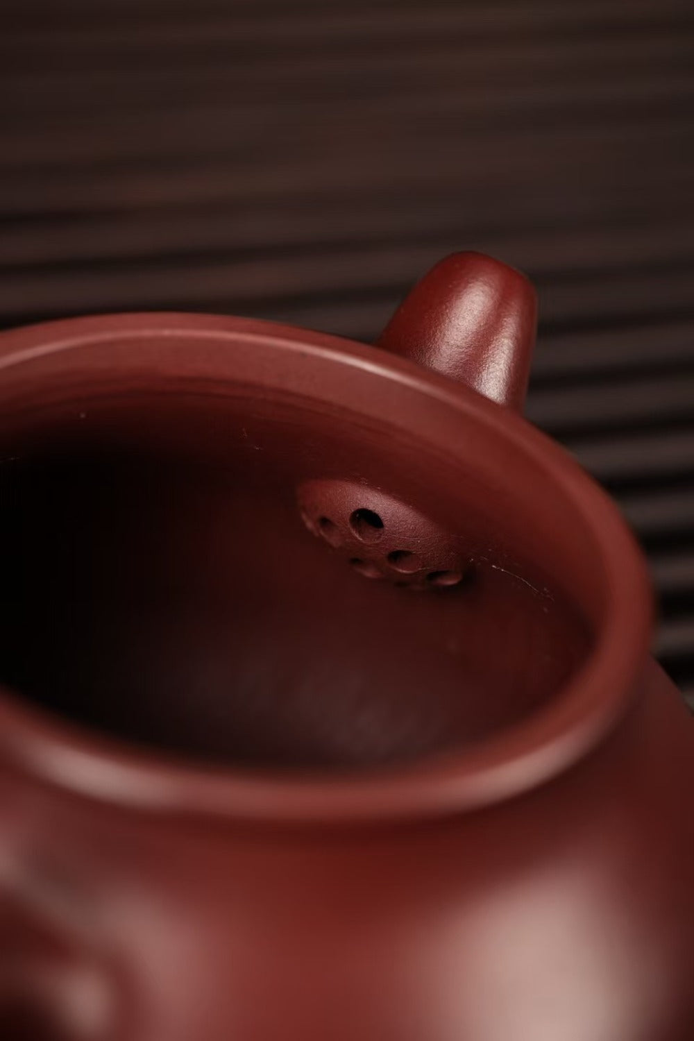 Close-up of a dark reddish-brown ceramic teapot showing the interior rim, spout, and drainage holes, set against a dark textured background.

