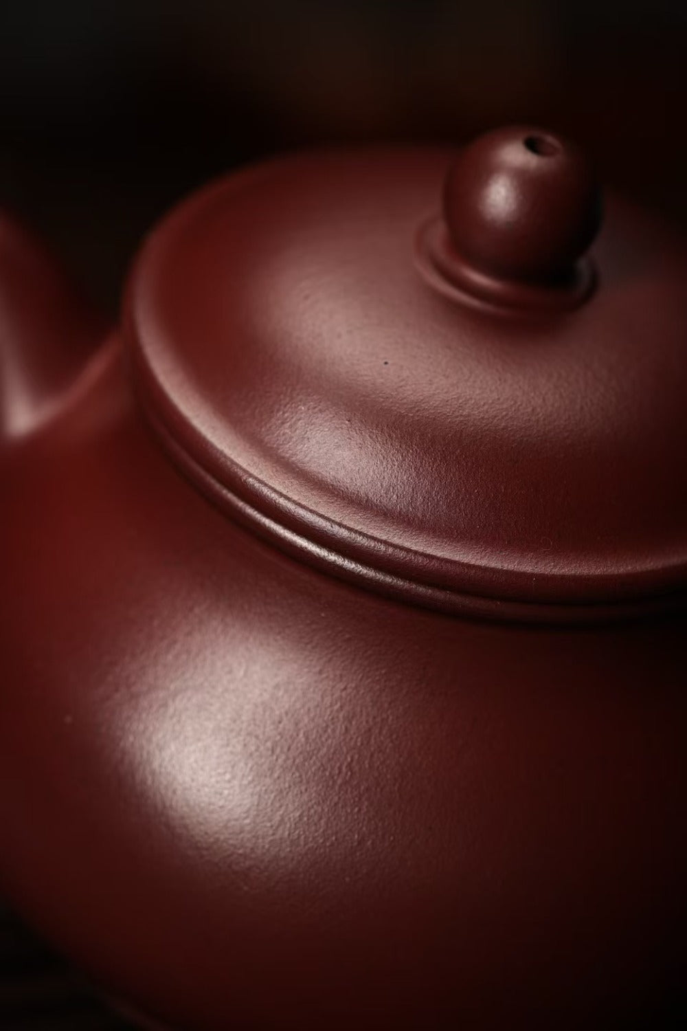 Close-up of a dark reddish-brown ceramic teapot with a smooth matte finish and a matching lid, photographed against a dark blurred background.

