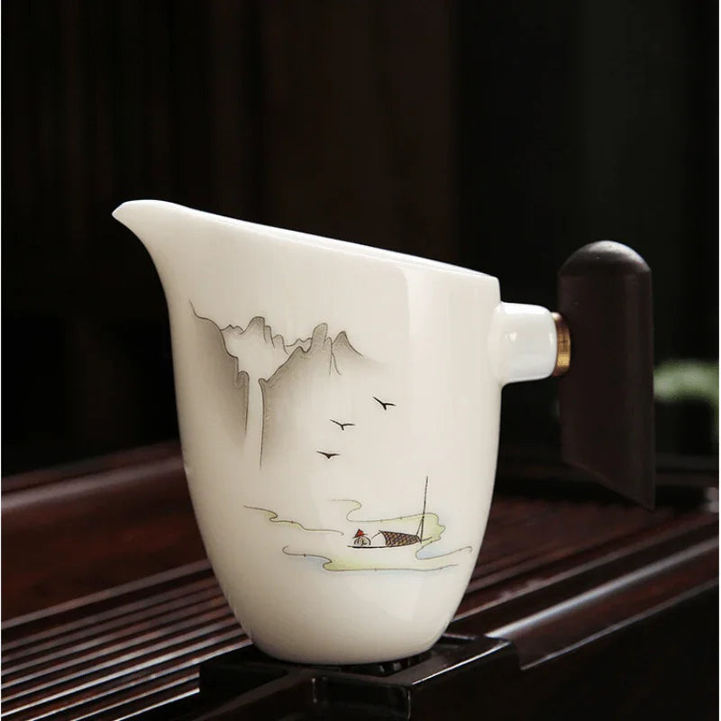A white ceramic pitcher with a hand-painted scenic design of mountains, birds, and a boat, resting on a dark wooden surface.

