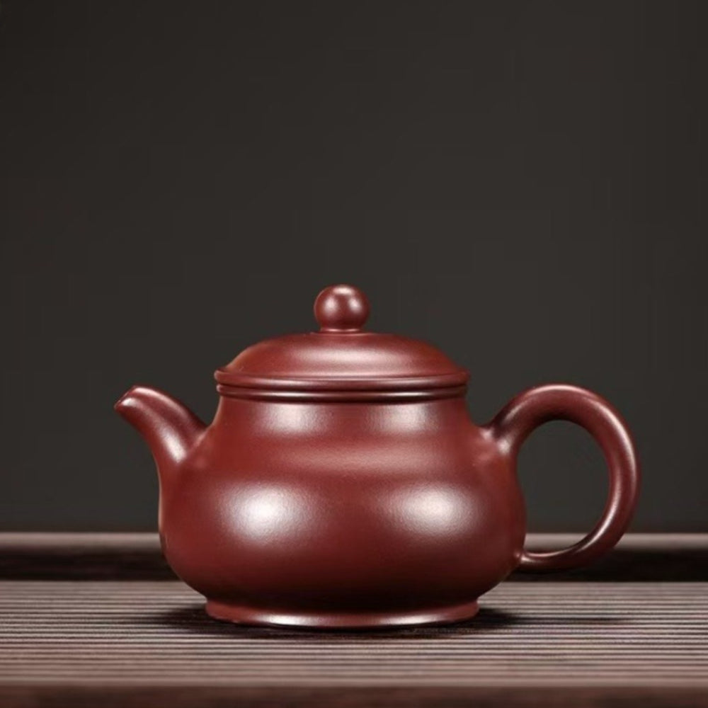 A classic Chinese Yixing Zisha teapot in a deep reddish-brown clay color, sitting on a dark wooden slatted surface against a dark gray background.

