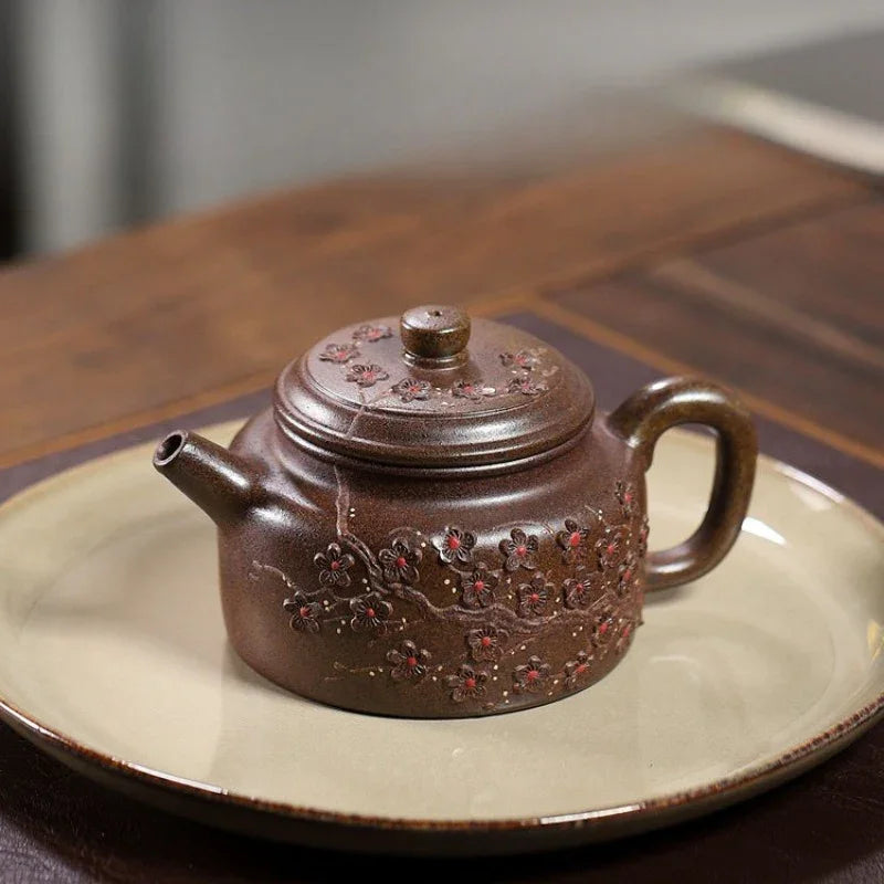 A brown ceramic teapot with embossed floral details, resting on a beige ceramic plate over a dark wooden table.

