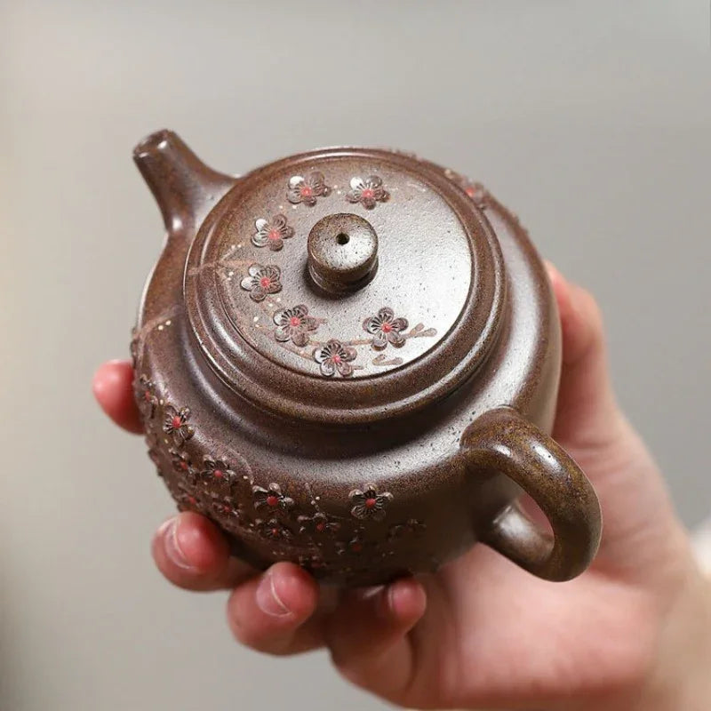 A hand holding a small brown ceramic teapot decorated with floral patterns, set against a plain light gray background.


