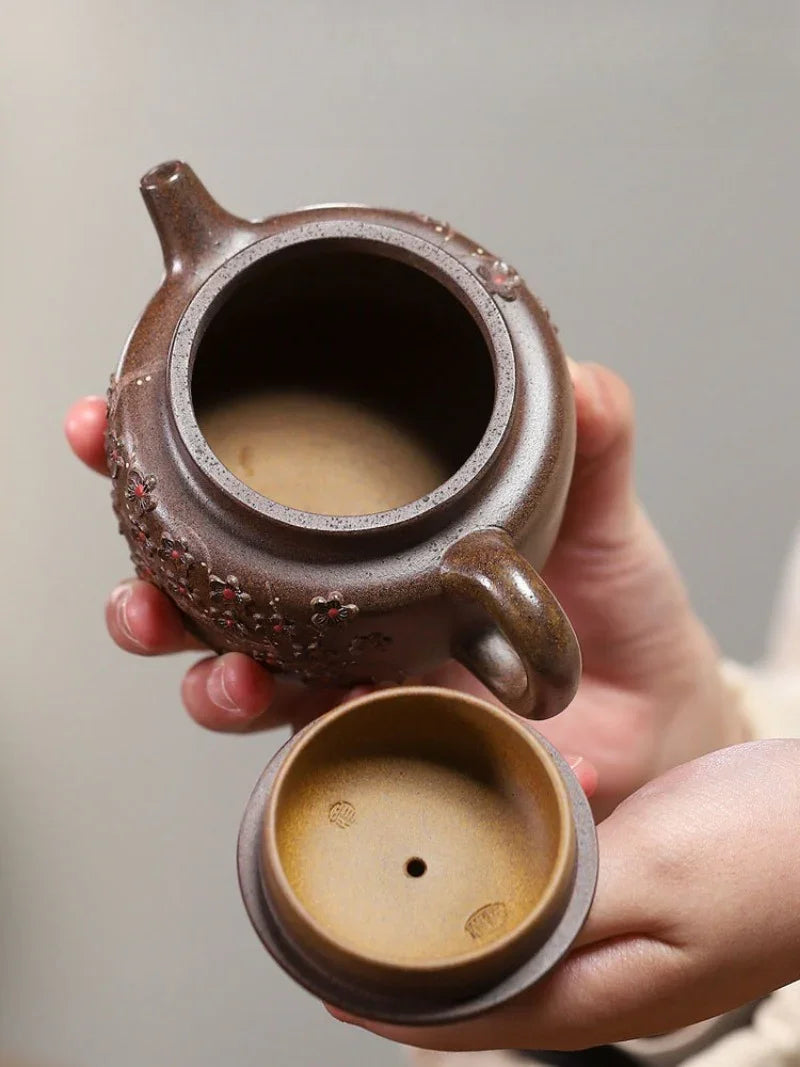 A close-up of a hand holding an open ceramic teapot, showing the brown exterior and light tan interior, with the lid resting below the spout against a soft gray background.

