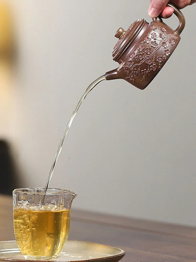 A hand holds a small brown ceramic teapot with embossed floral detailing, pouring tea from a clear glass teacup resting on a plate.

