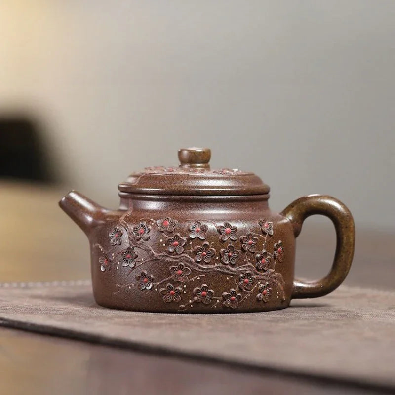 A brown ceramic Yixing teapot with embossed floral patterns and small red accents, resting on a textured brown mat against a blurred gray background.

