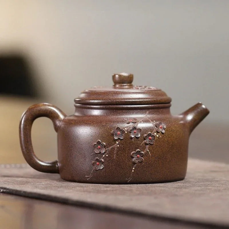 A brown ceramic teapot with a lid, featuring embossed cherry blossom flowers on the side, resting on a textured mat.

