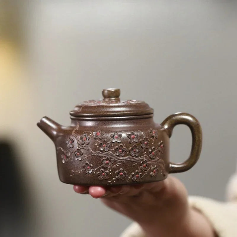 A hand holding a small brown ceramic teapot with embossed floral patterns, against a softly blurred neutral background.

