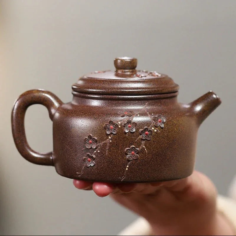 A brown ceramic teapot with floral engravings, held in a person's hand against a neutral gray background.

