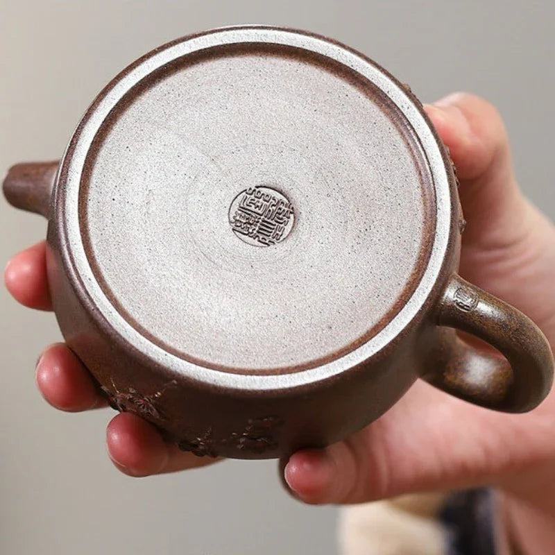 A close-up of a hand holding the bottom of a brown ceramic yixing tea pot, showing the textured clay surface and a circular maker's seal stamp in the center.

