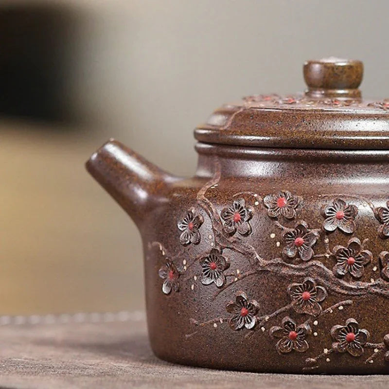 A close-up of a brown ceramic teapot decorated with embossed cherry blossom branches and red-painted flower centers, shown on a neutral surface with a softly blurred background.

