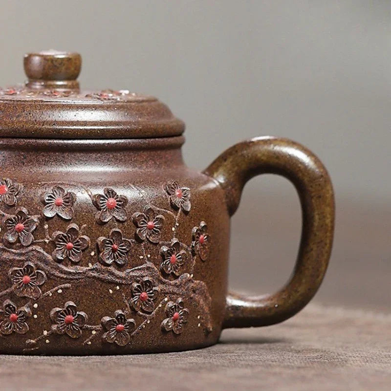 A close-up of a brown ceramic teapot with embossed cherry blossom branches and red-pink flower centers, set against a neutral gray background.


