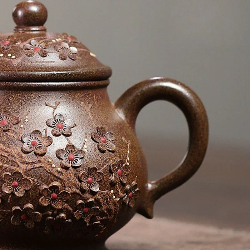 A close-up of a brown ceramic teapot with embossed cherry blossom flowers and small red centers, set against a soft gray background.

