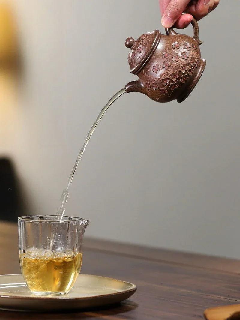 A hand holds a brown ceramic teapot pouring tea into a clear glass cup on a saucer, set on a wooden table against a softly blurred background.

