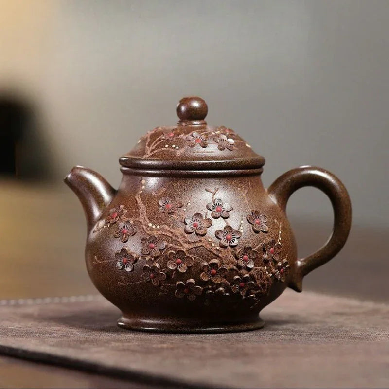 A brown ceramic teapot with a lid, decorated with embossed cherry blossom branches and pink flowers, resting on a dark textured mat against a blurred gray background.

