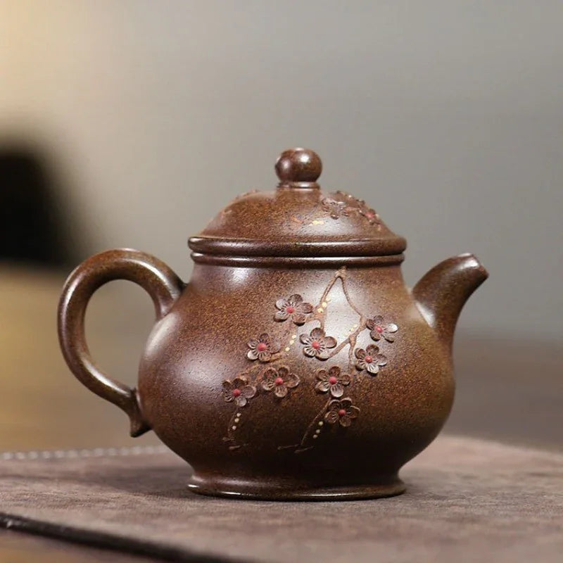 A brown ceramic teapot with a lid, featuring hand-painted cherry blossom details on the front, resting on a dark wooden surface with a blurred background.

