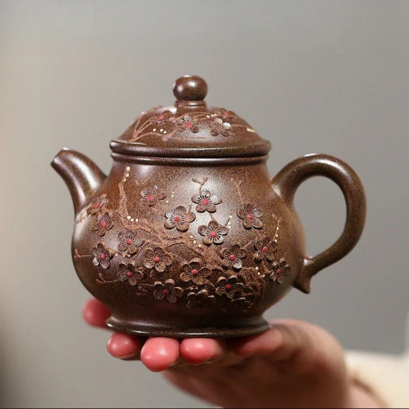 A hand holding a brown ceramic teapot decorated with embossed floral patterns and small red accents, set against a plain gray background.

