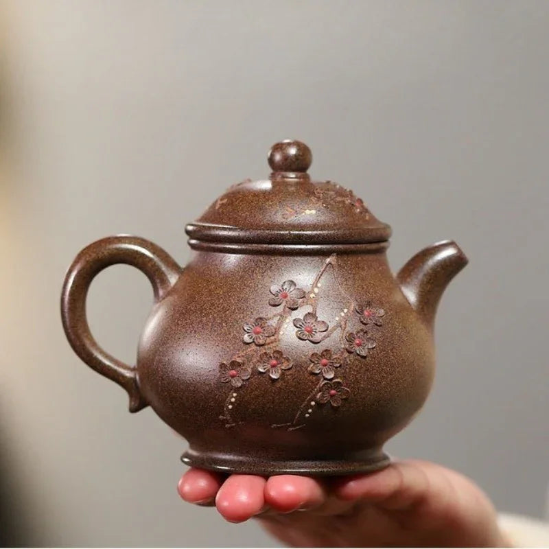 A brown ceramic teapot with embossed cherry blossom details, held in a person's hand against a soft gray background.

